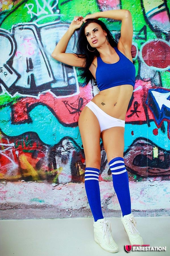 Jasmine Jae in sporty outfit and socks and trainers doing a strip against a graffiti wall